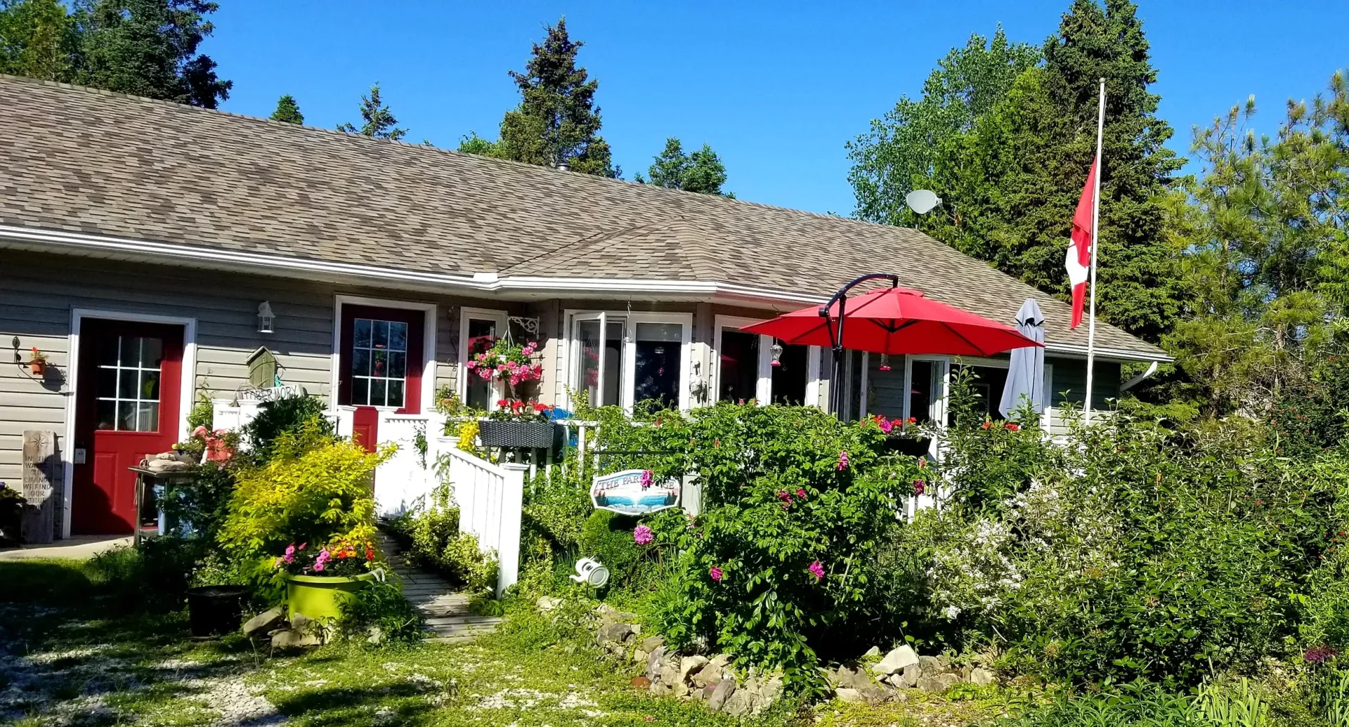A cottage with a garden, red umbrellas, and a Canadian flag under a clear blue sky