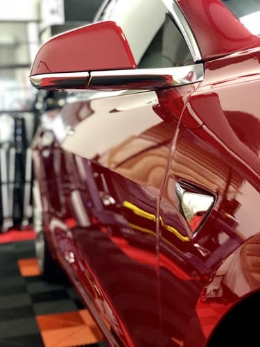 Red Tesla car, shiny exterior, side mirror, chrome trim, on a patterned floor.