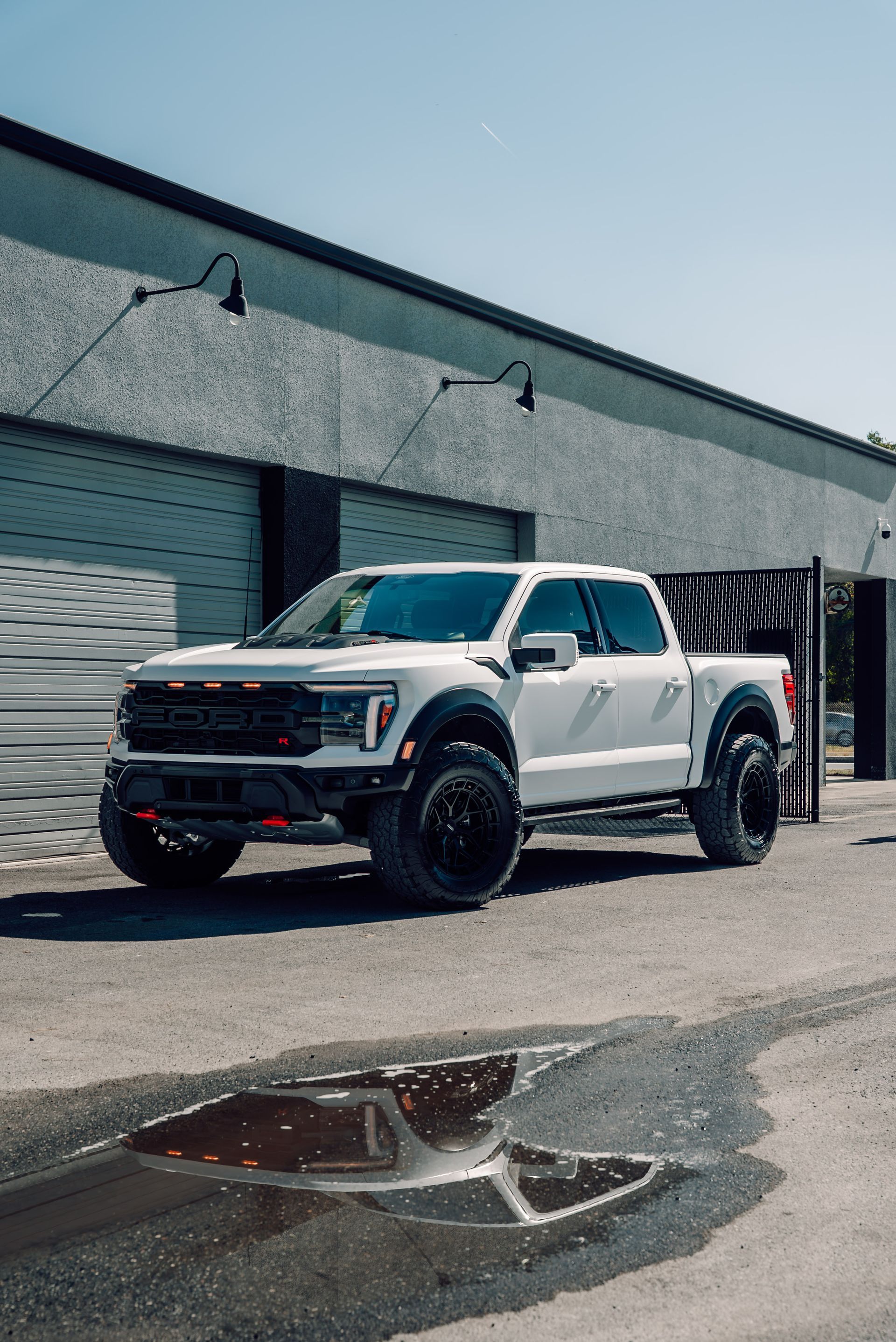 White Ford F-150 Raptor truck parked on a street with a garage in the background. Black wheels. Sunny day.
