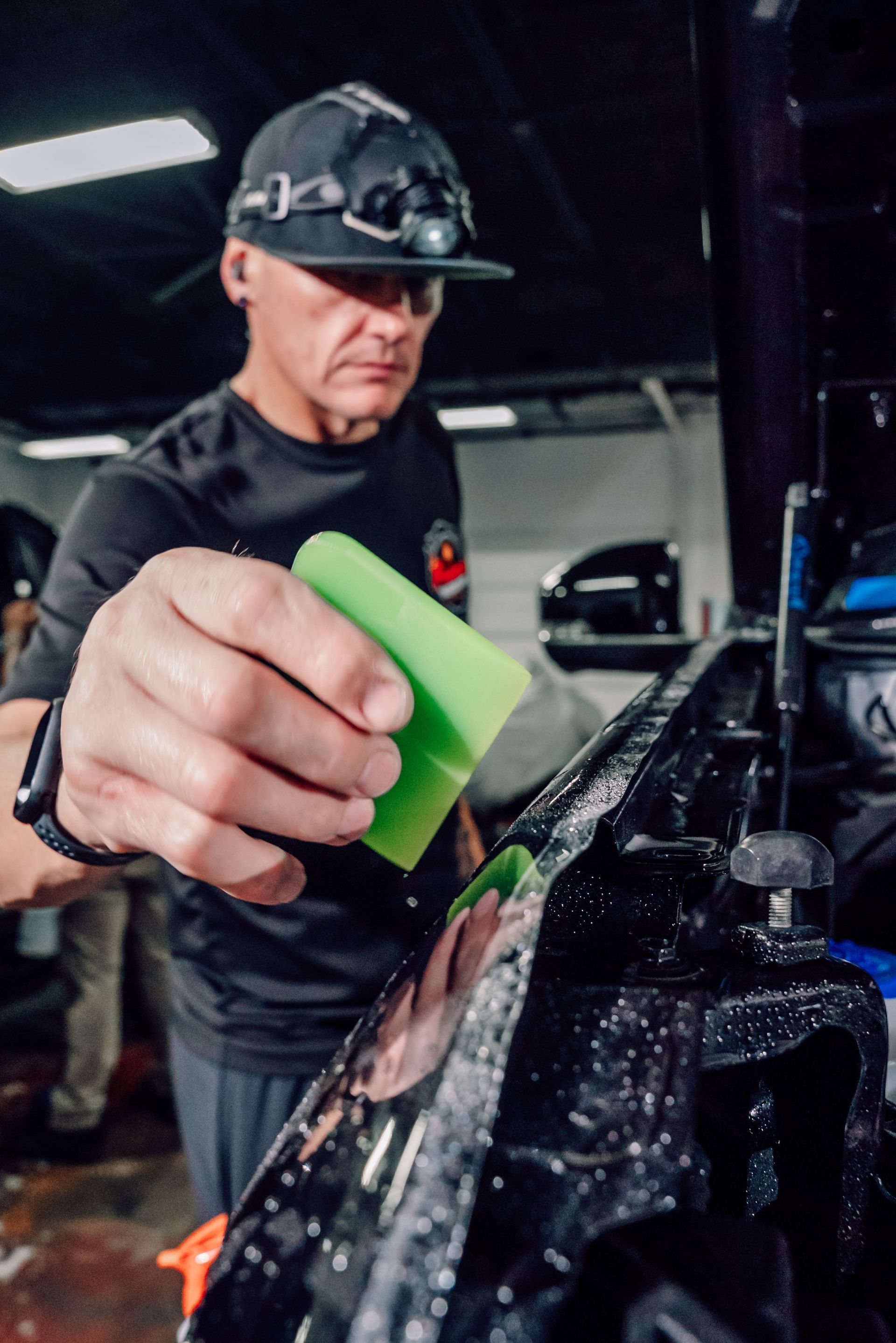 A person wearing a headlamp uses a green squeegee to apply a protective film to a wet, black vehicle panel.
