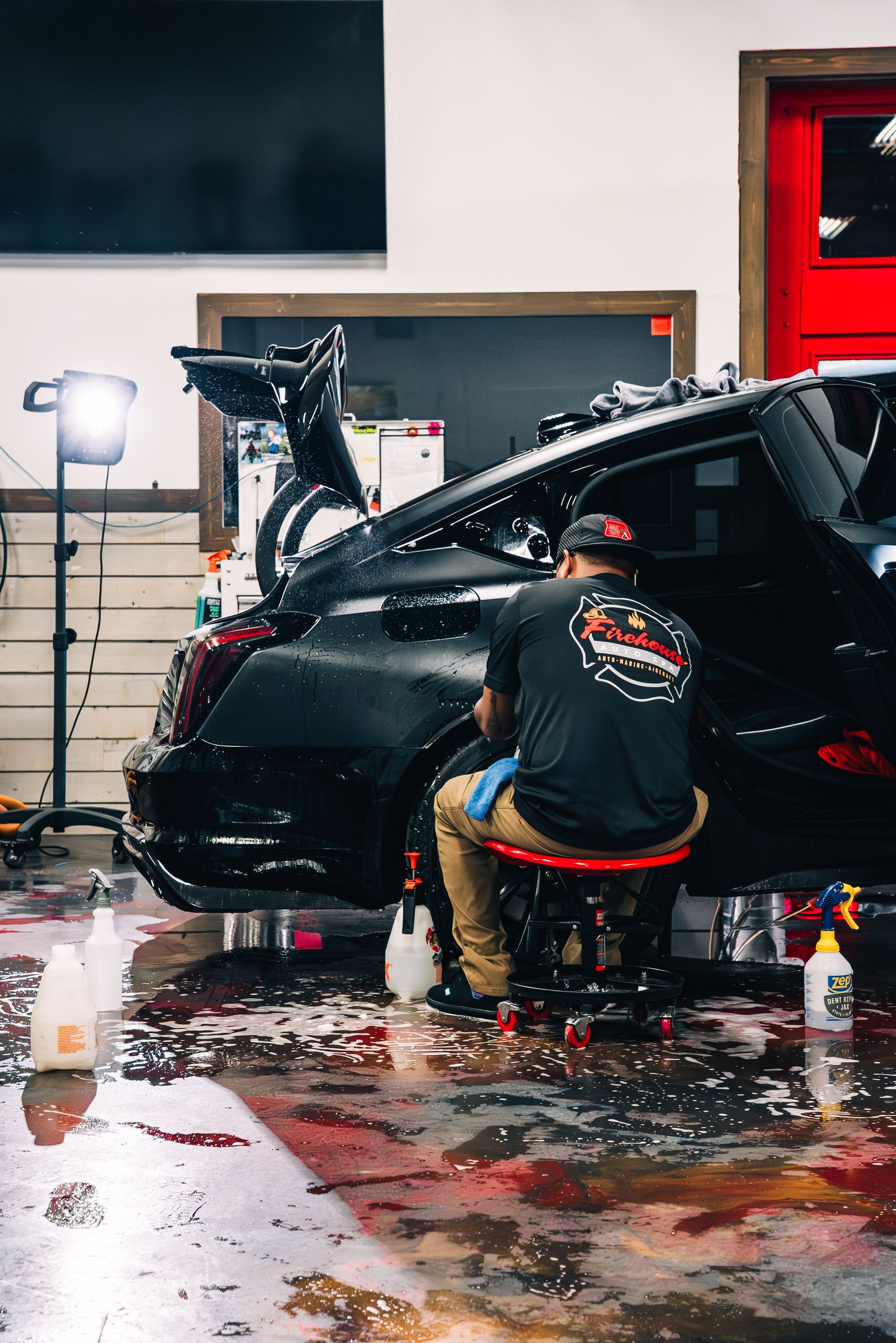 A worker sits on a rolling stool, detailing the rear wheel area of a black car in a brightly lit shop with a wet floor.