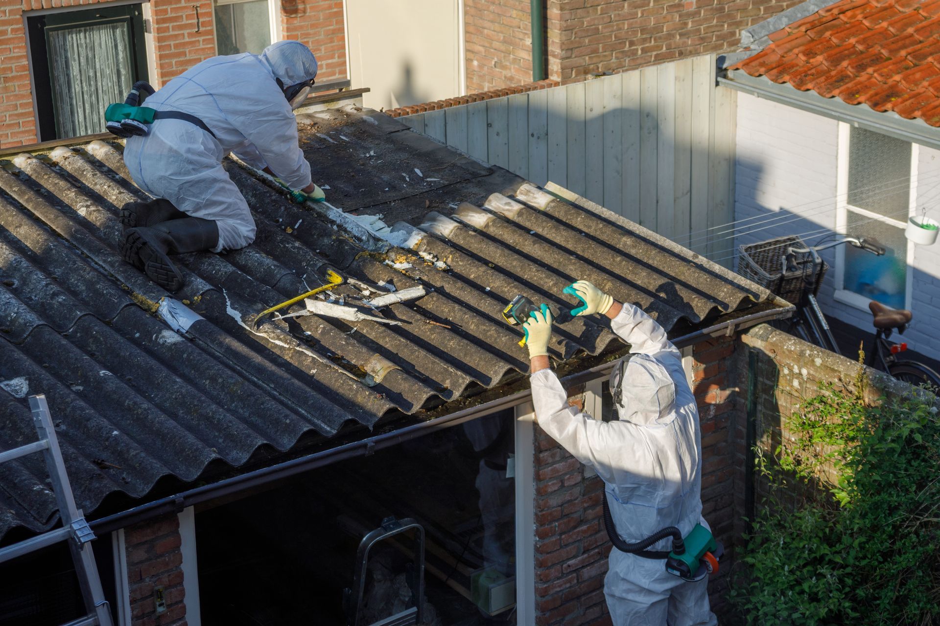 Two workers in protective suits removing asbestos roof panels outdoors.