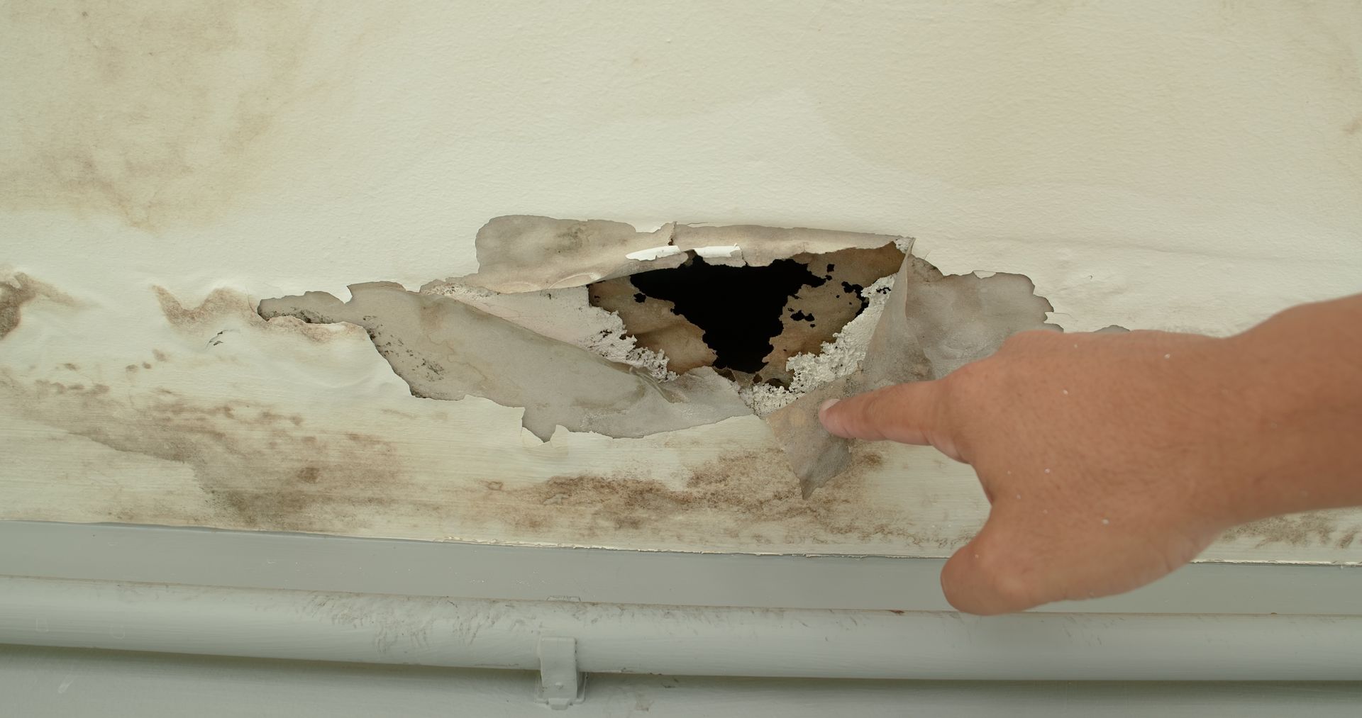 Hand pointing at damaged ceiling with a hole, surrounded by water stains and peeling paint.