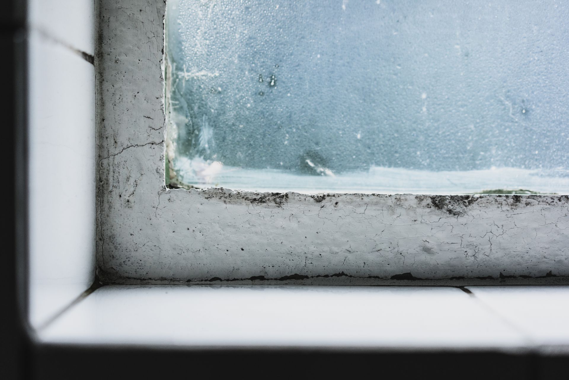 Close-up of a window corner with frosted glass, surrounded by a worn, white frame and tiled wall.