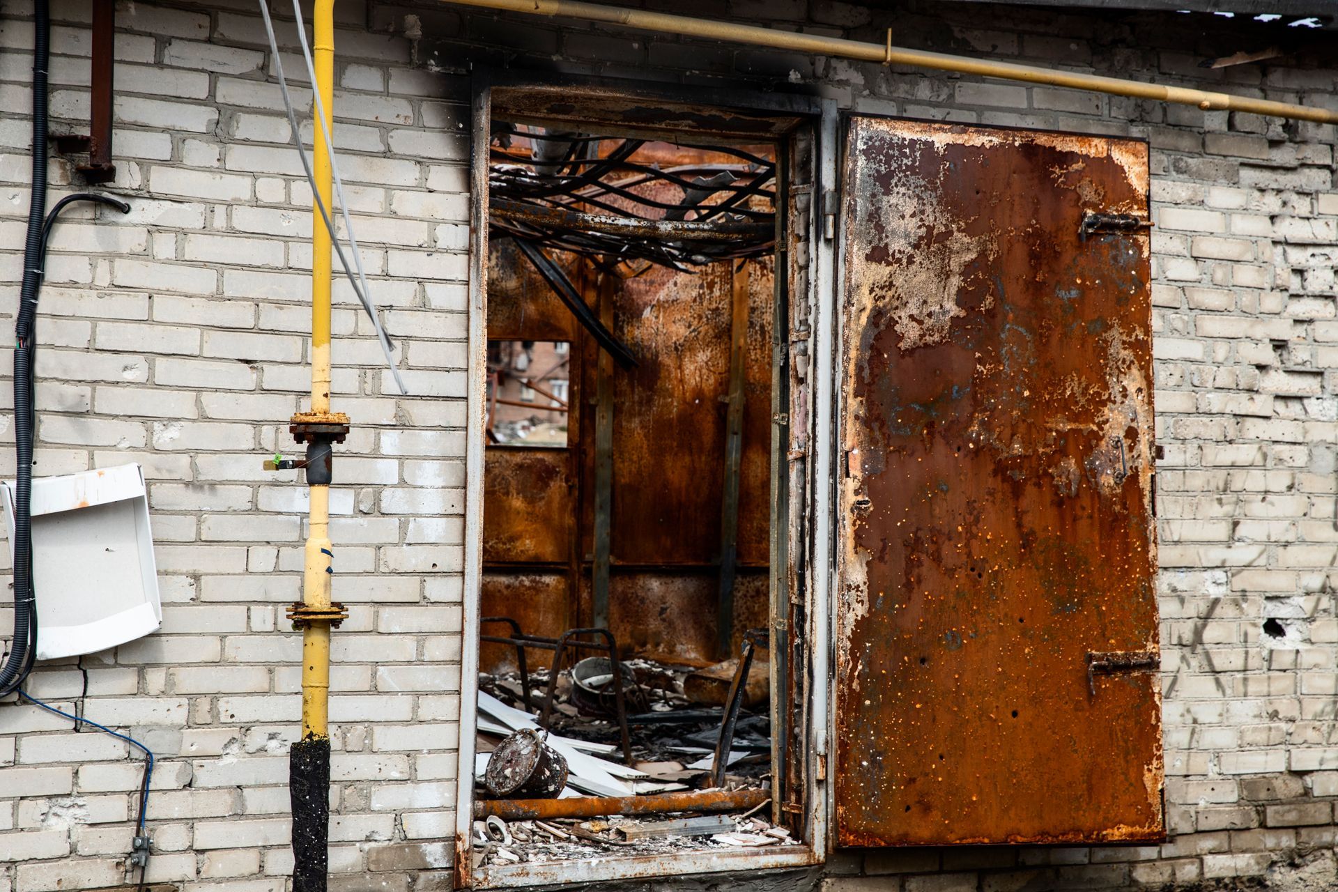 a rusty and burnt door due to fire is open on the side of a brick building .