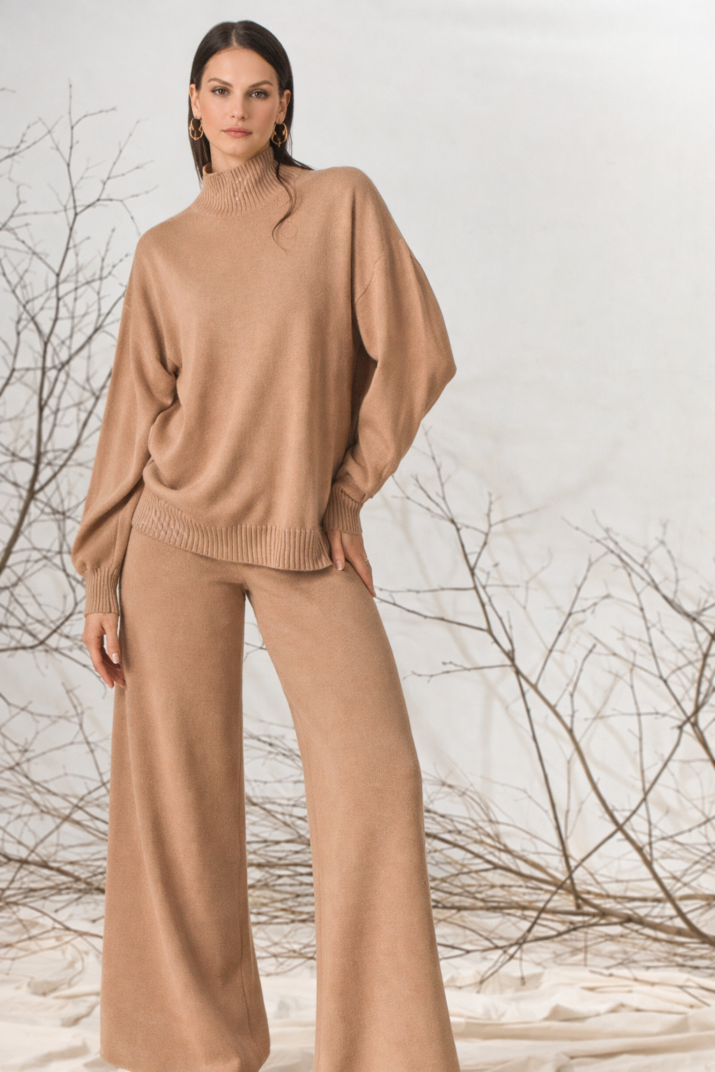 Donna con maglione e pantaloni color cuoio, in piedi davanti a rami spogli.