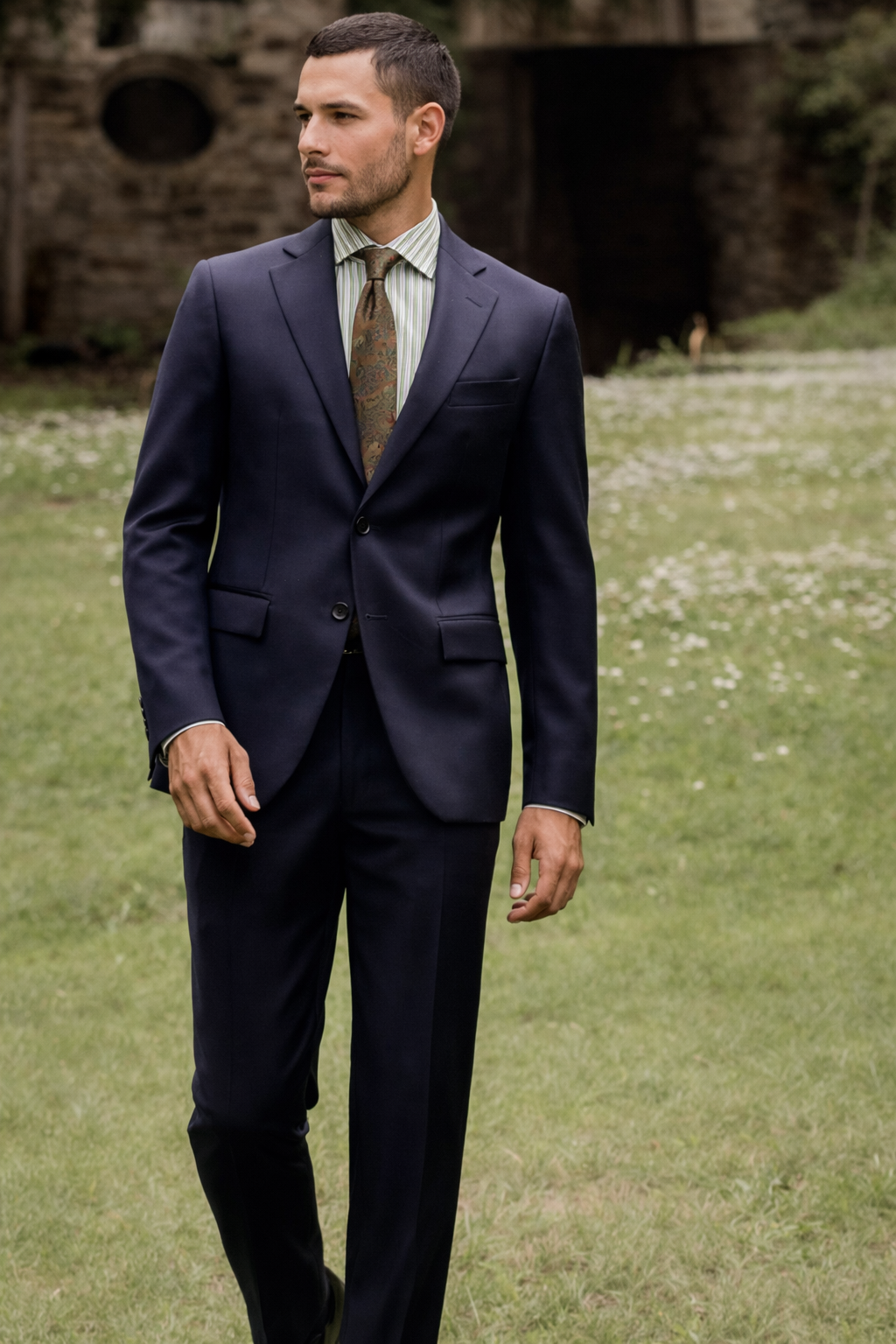 Un uomo in giacca e cravatta blu navy cammina in un prato vicino a un edificio in pietra.