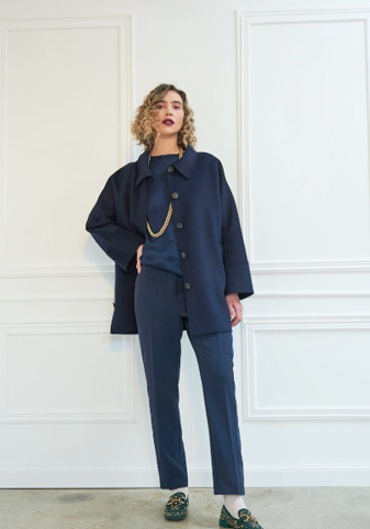 Donna in abito blu navy con giacca e pantaloni, catena d'oro e mocassini. In piedi davanti a un muro bianco.