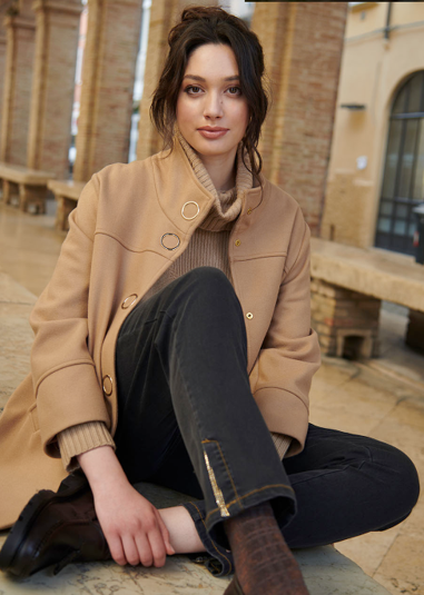 Una donna con un cappotto marrone chiaro e jeans è seduta all'aperto.