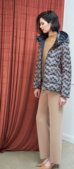 Donna con giacca stampata, maglione marrone chiaro e pantaloni. In piedi vicino a una tenda rossa.