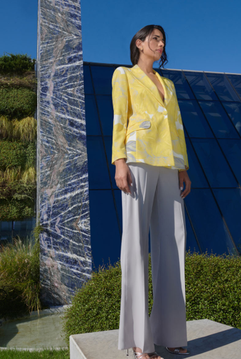 Donna con giacca gialla e pantaloni grigi, in piedi all'aperto, che guarda di lato.