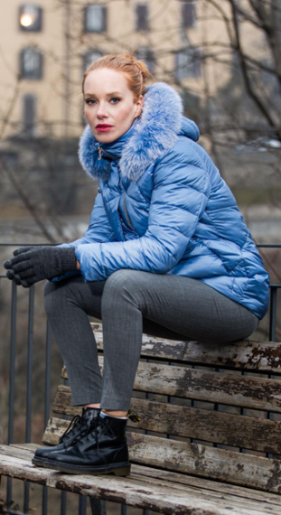 Donna con piumino blu e cappuccio di pelliccia, seduta su una panchina, con guanti e stivali.
