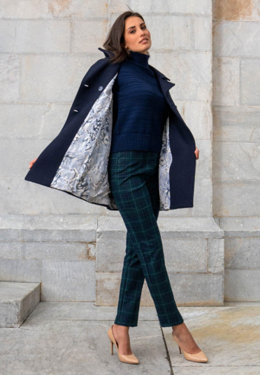 Donna con cappotto e maglione blu navy, pantaloni a quadri verdi e tacchi; posa contro un muro di pietra.