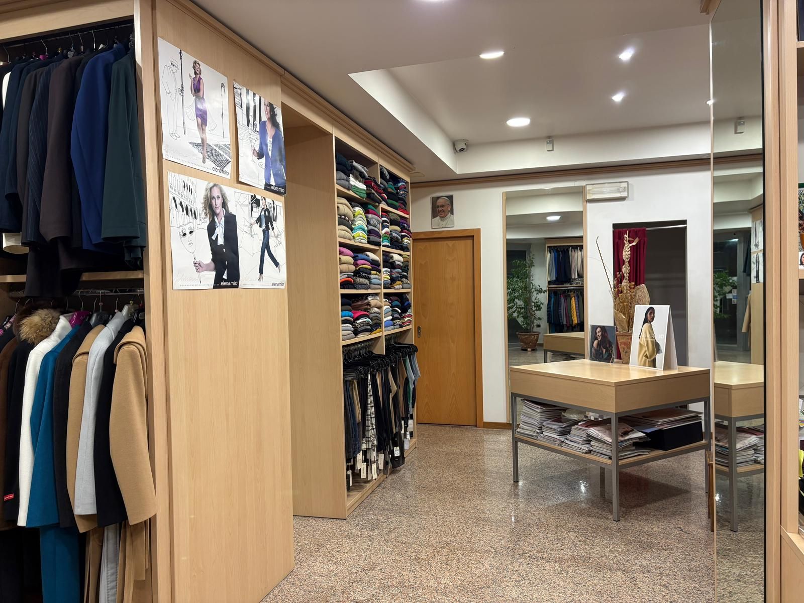 Interno di un negozio di abbigliamento con scaffali pieni di vestiti e tavoli espositivi,
