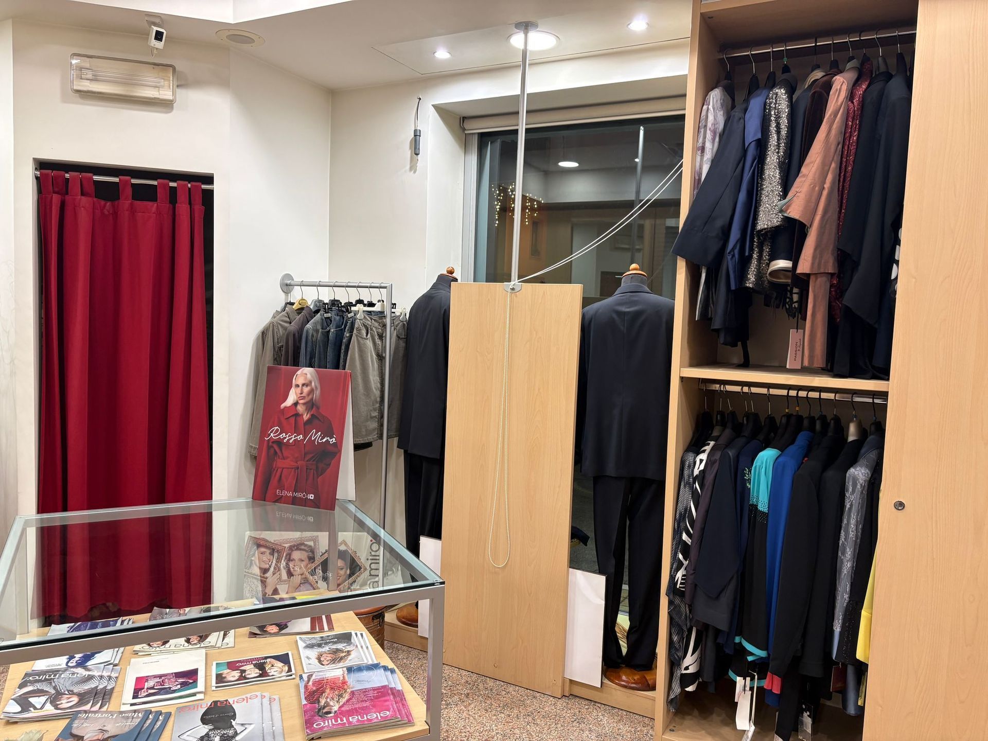 Interno di un negozio di abbigliamento con scaffali di vestiti, manichini, vetrina e tenda rossa.