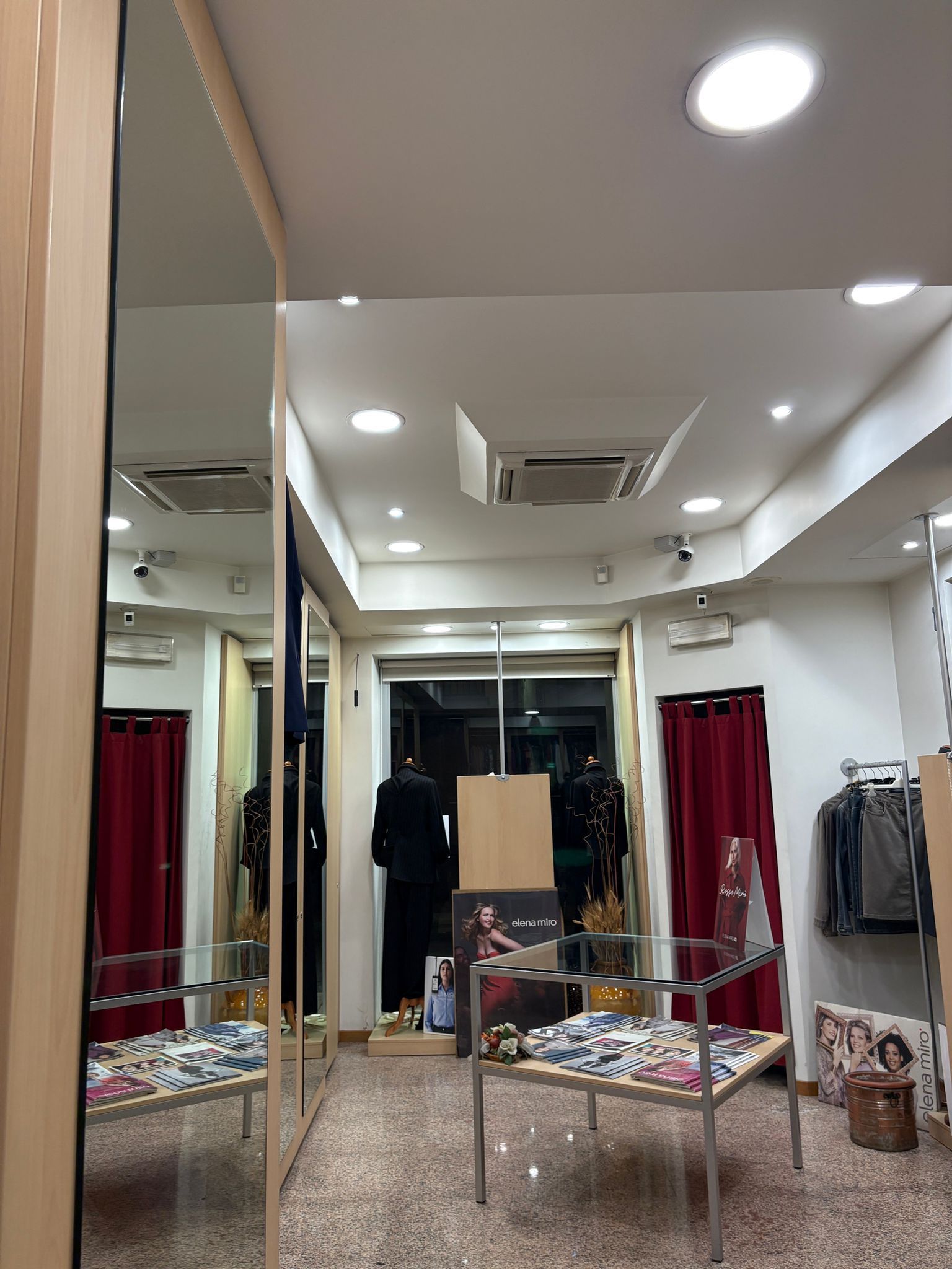 Interno di un negozio di abbigliamento, con uno specchio che riflette manichini, sedie e abiti esposti.