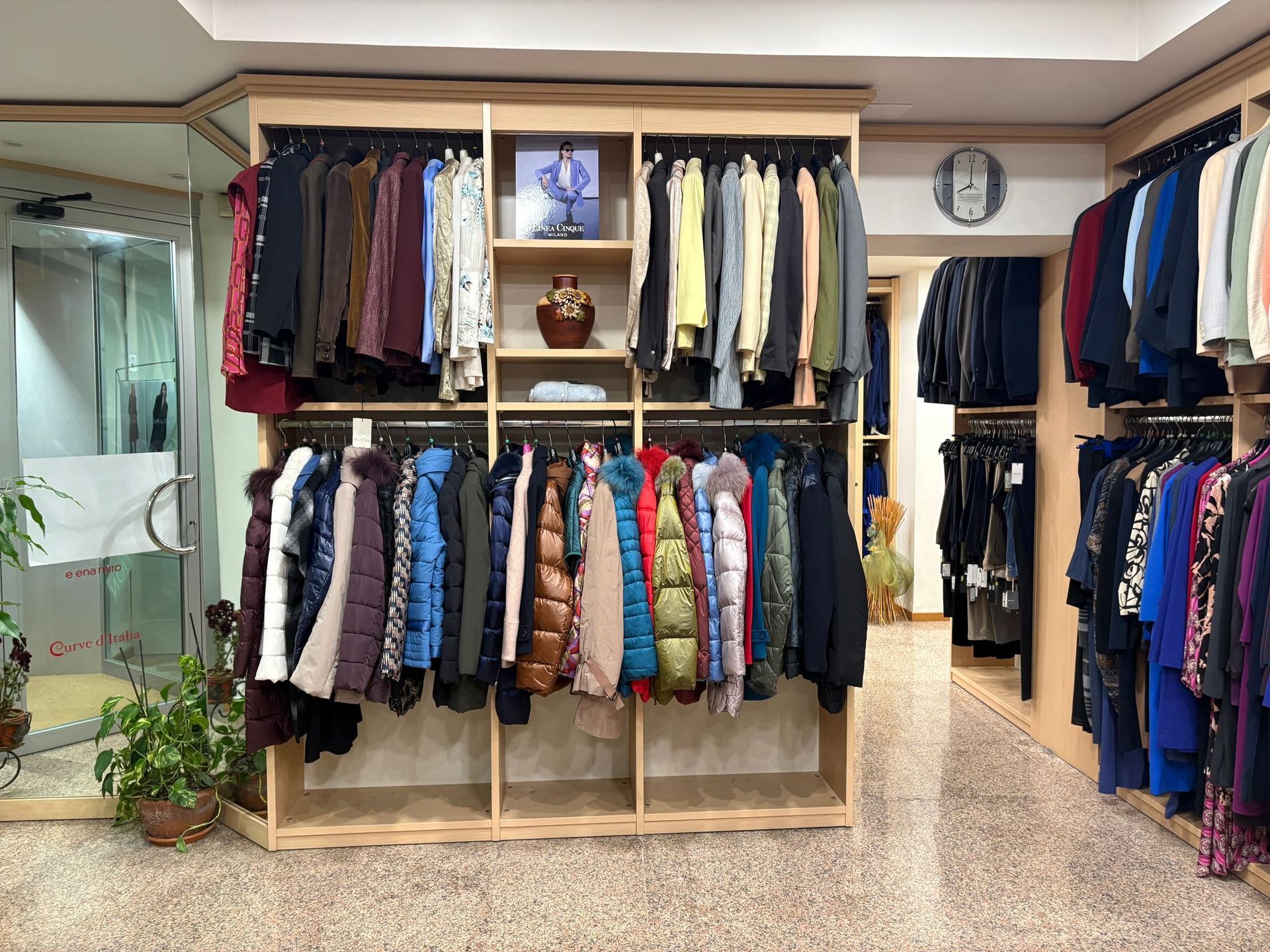 Interno di un negozio di abbigliamento con scaffali pieni di giacche, camicie e vestiti, illuminati da luci soffuse.