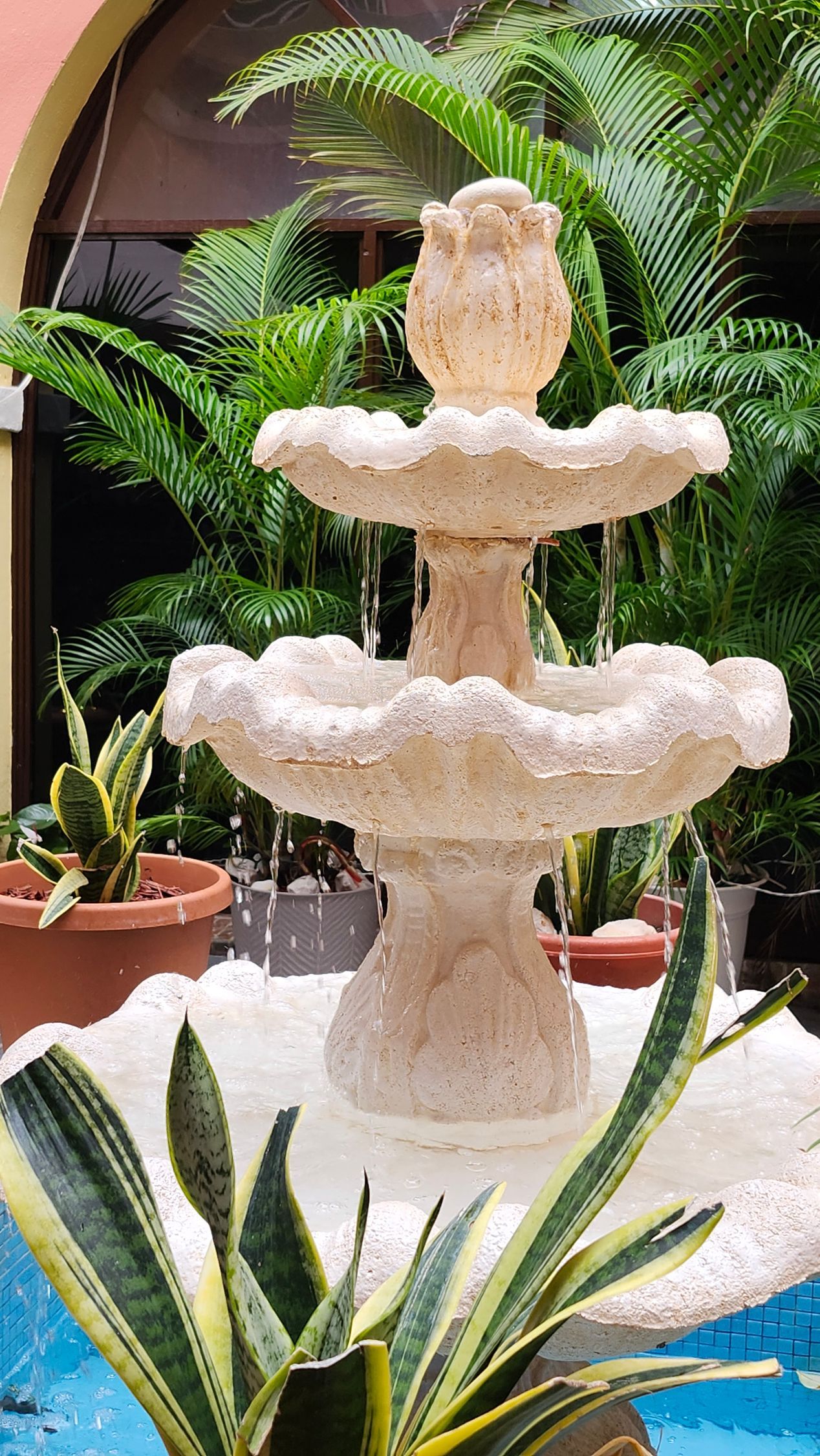 Three-tiered white fountain in a lush courtyard with potted plants and a blue pool edge