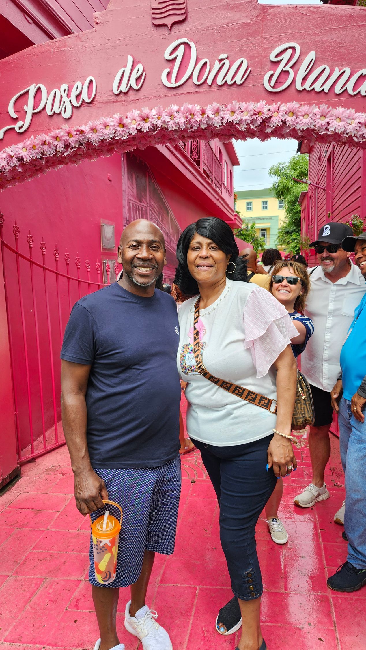 Two people pose on a pink path under a “Rosa de…” arch, holding drinks in a festive outdoor setting.