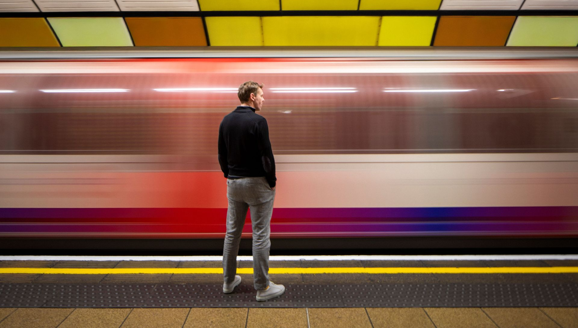 Person stands on a subway platform as a blurred train rushes past under colorful ceiling panels