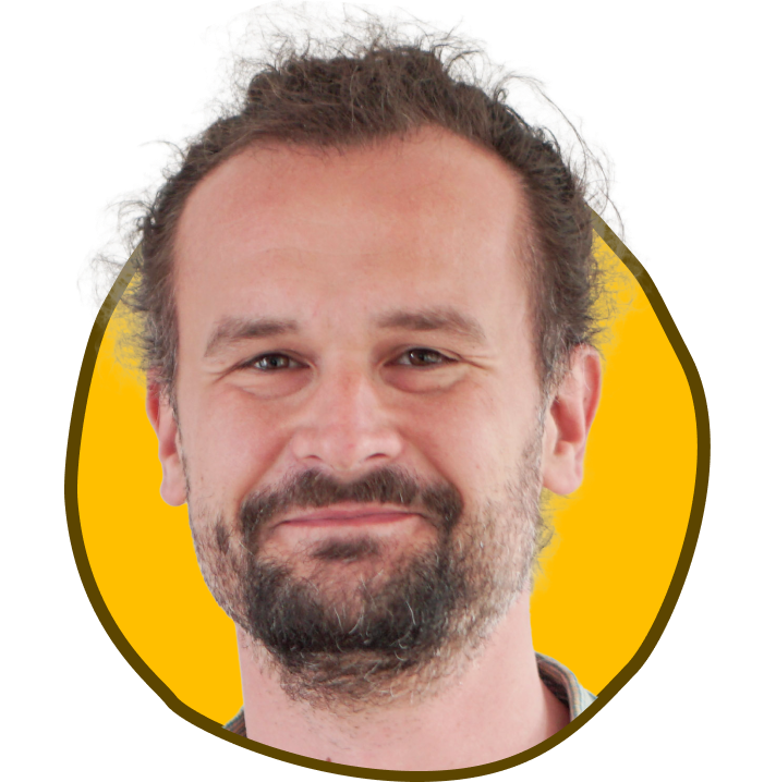 Mand med krøllet brunt hår og skæg smiler, indrammet i en gul cirkel.