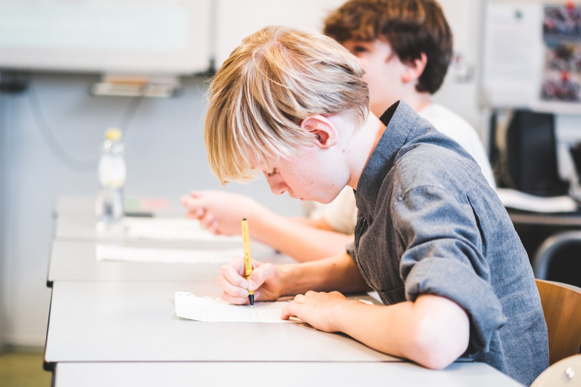 A boy is sitting at a desk in a classroom writing on a piece of paper.
