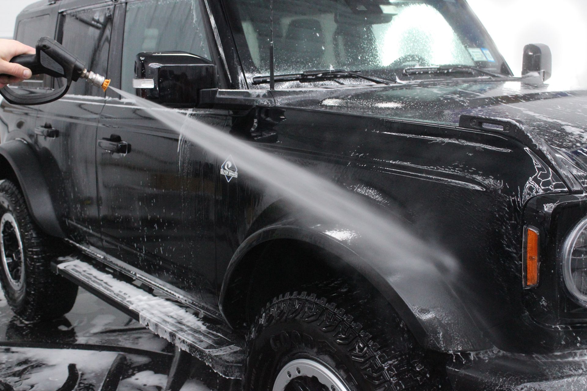 A person power-washing a black SUV, spraying water onto the front fender and side.