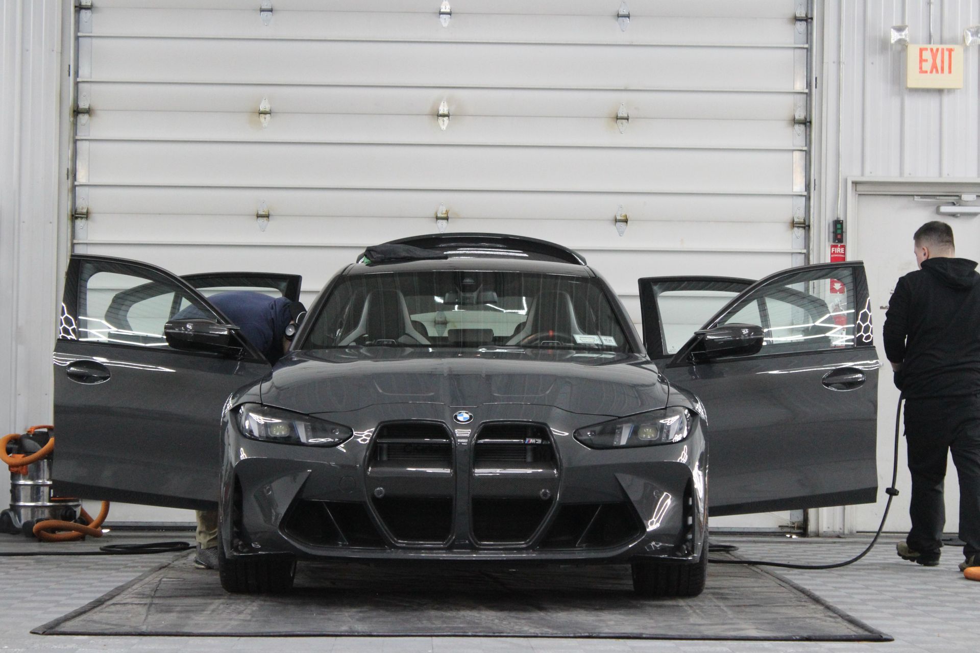 A charcoal BMW in a garage with open doors as someone works on the interior and another person stands nearby.