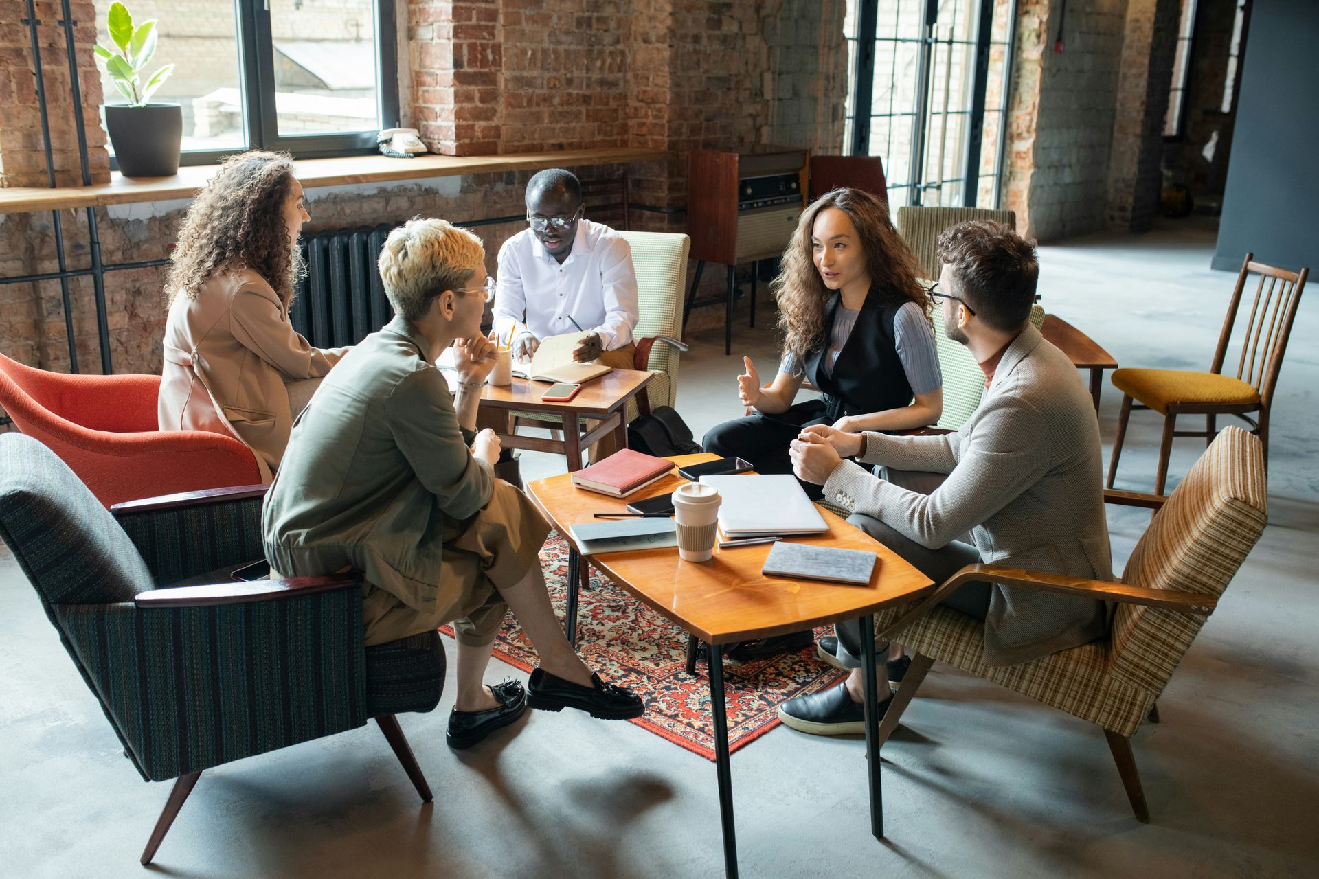Group of people in casual wear meeting in a room with exposed brick and furniture, discussing at a table.