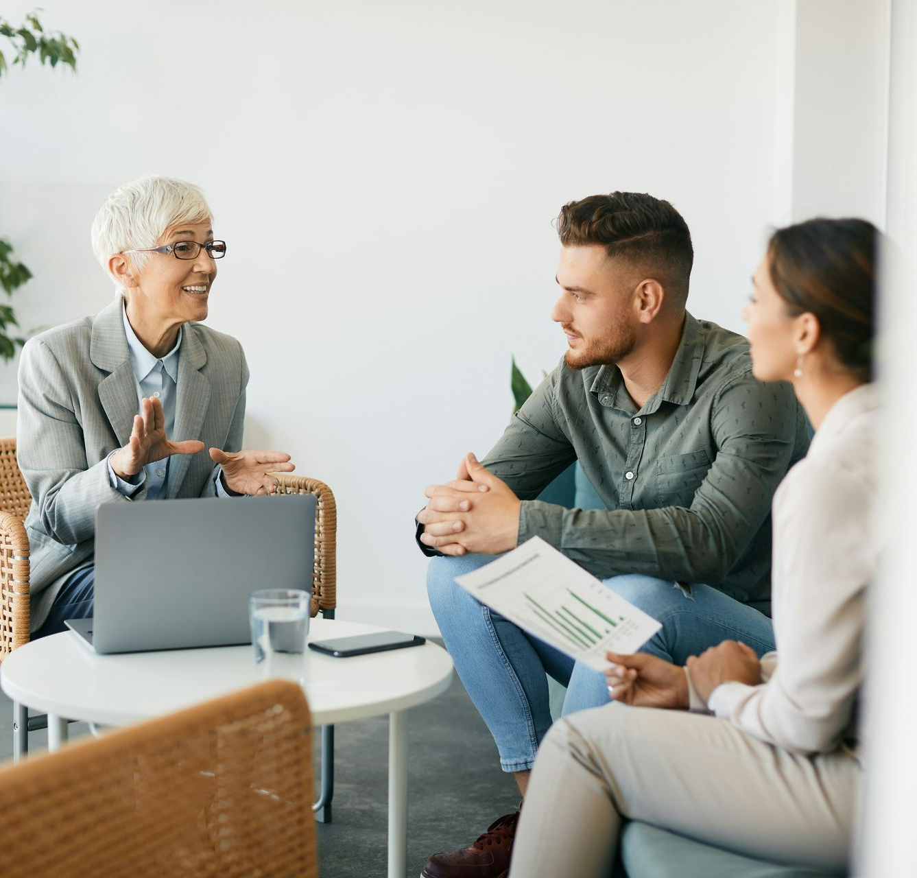 Woman in blazer talking to a couple in a meeting, laptop and documents on the table.