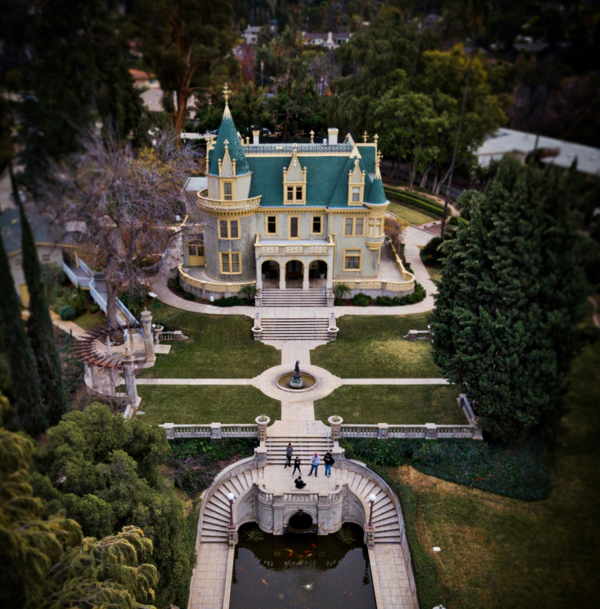 Aerial view of a Victorian mansion with green roof, formal garden, and people walking on a walkway.
