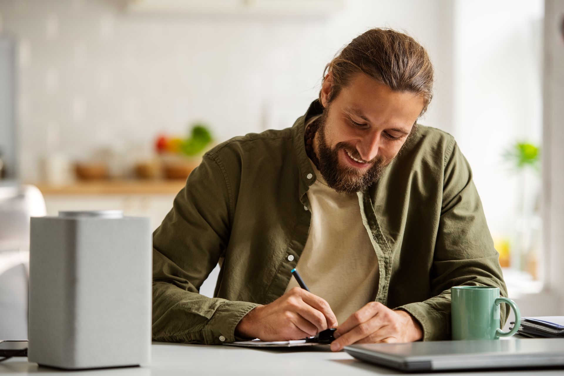 Man with beard writing at a table; mug, speaker, and laptop nearby. Kitchen setting, smiling.