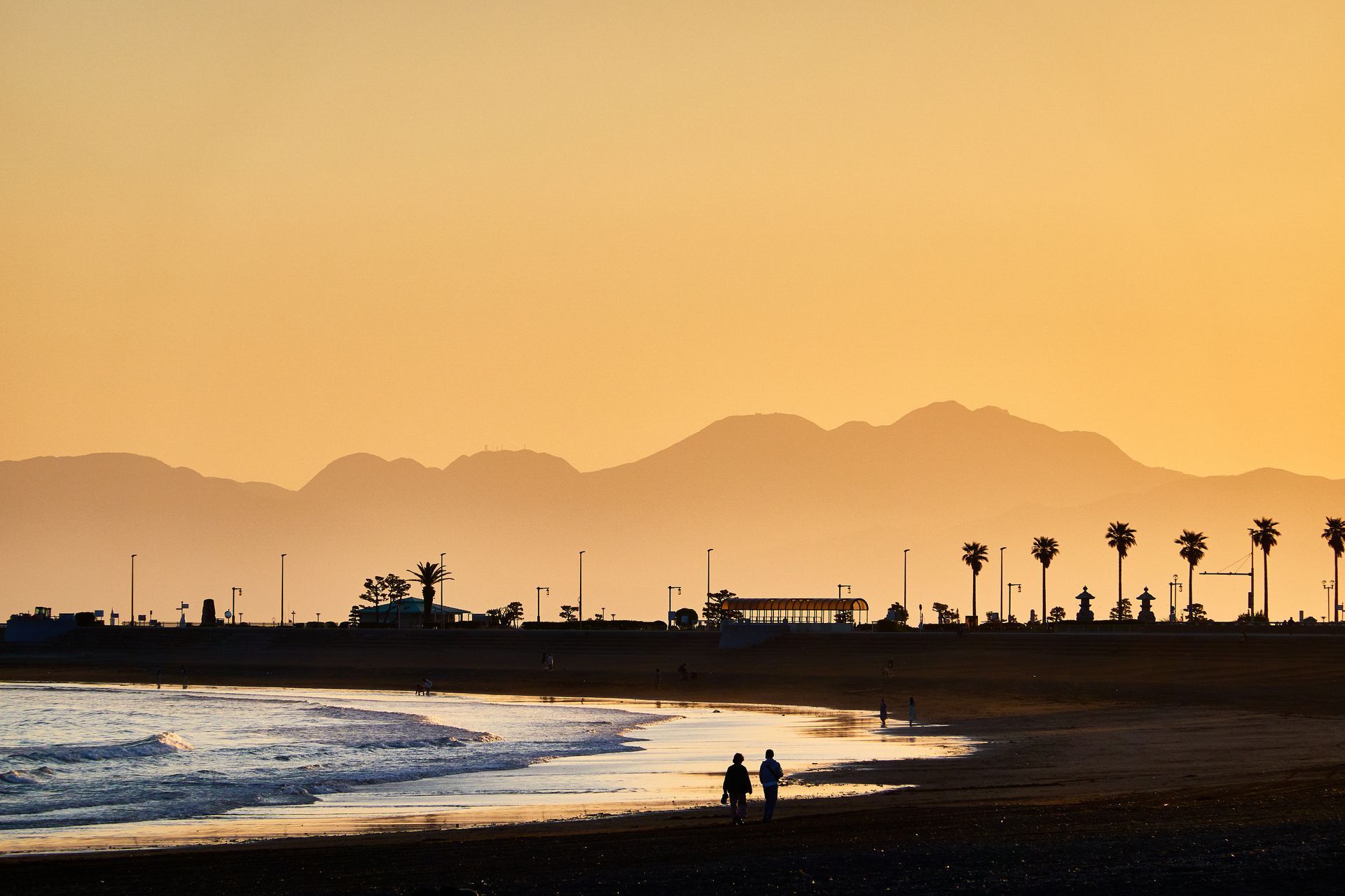 Beach at sunset with silhouetted figures, palm trees, and mountains in the distance.