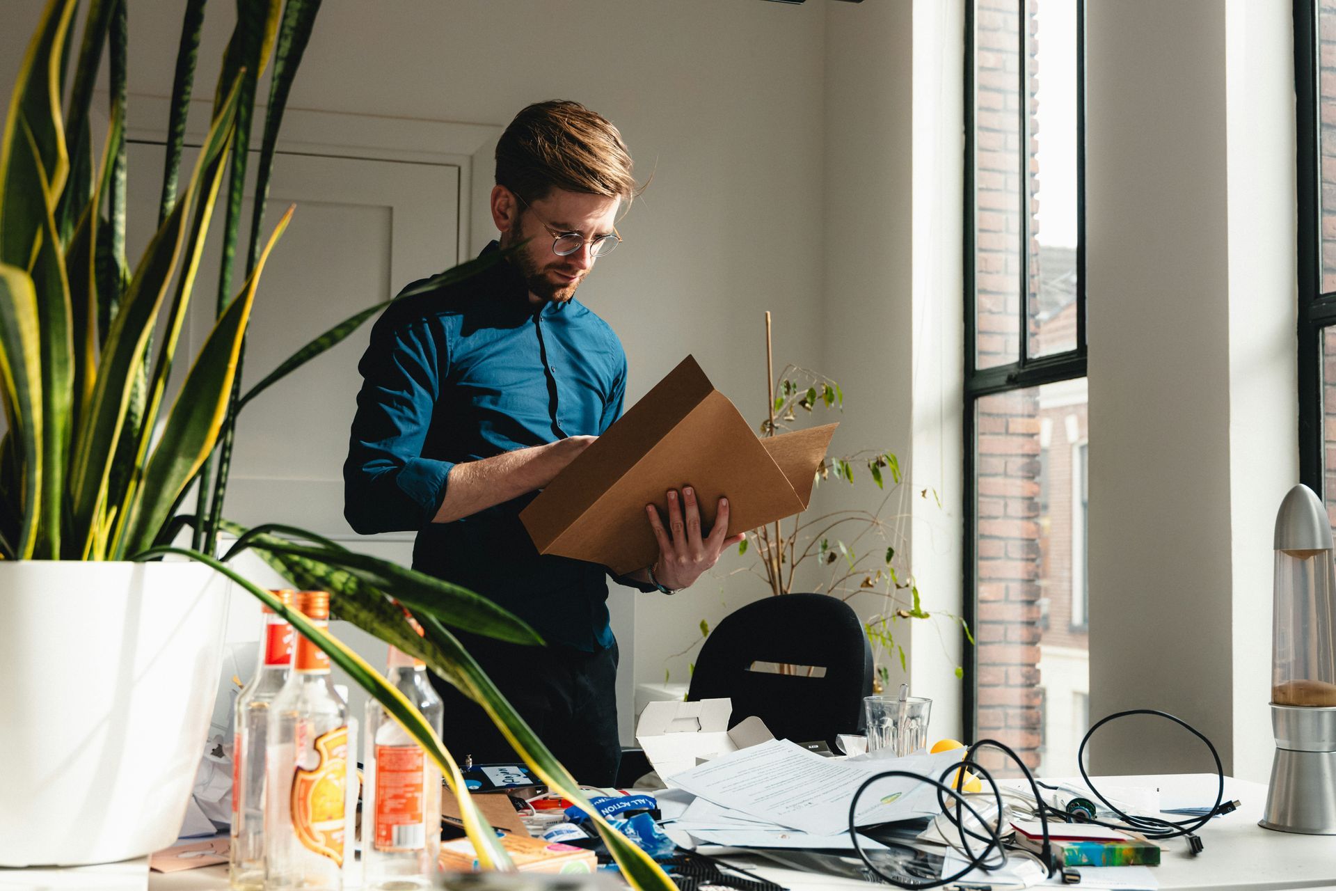 Man in glasses reads a cardboard file in a bright office, with a large window and potted plants.