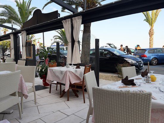 Ristorante all'aperto con tavoli bianchi, sedie e tettoia, vicino a una strada con auto e palme.