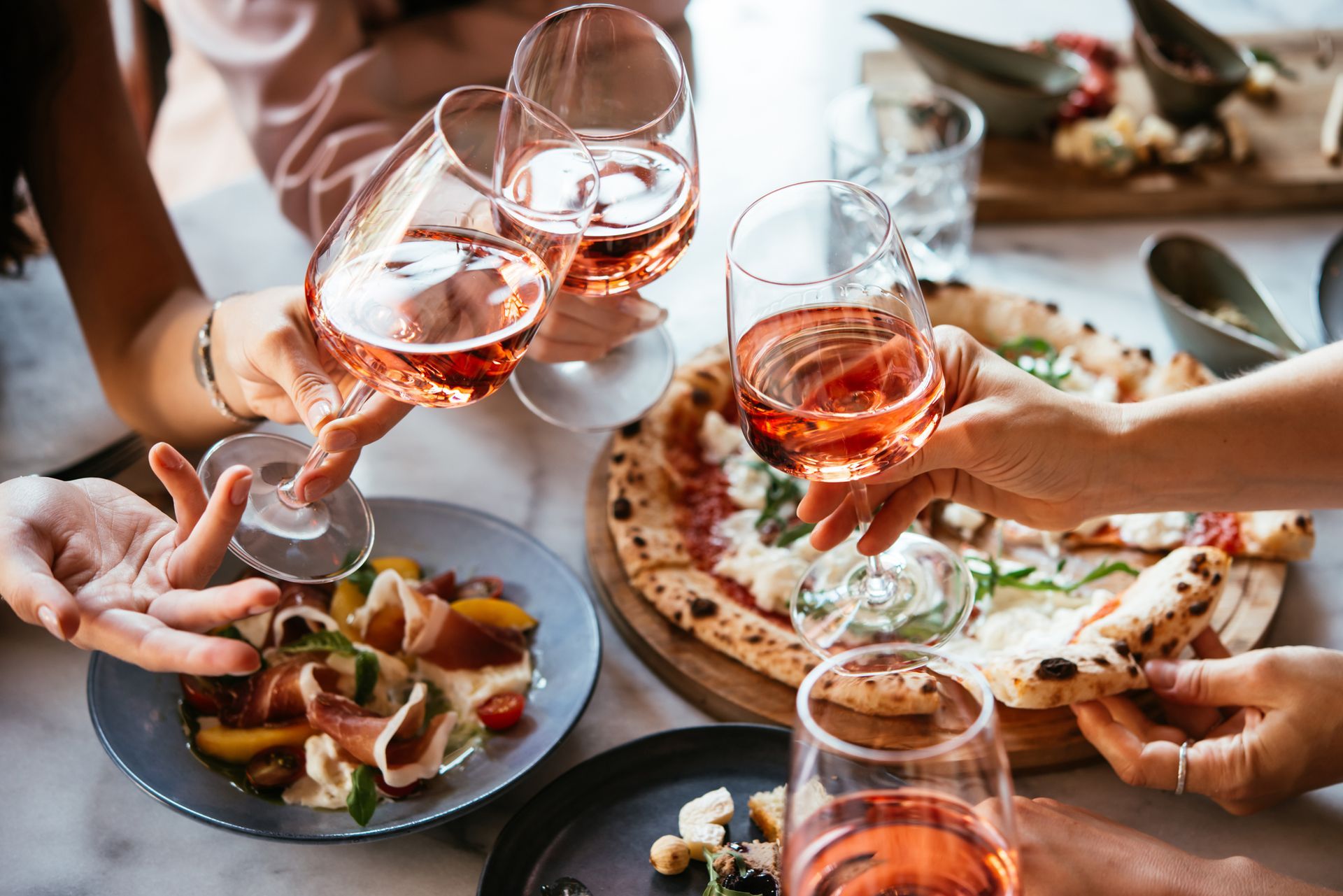 Persone che brindano a un tavolo con vino rosato, pizza e stuzzichini.