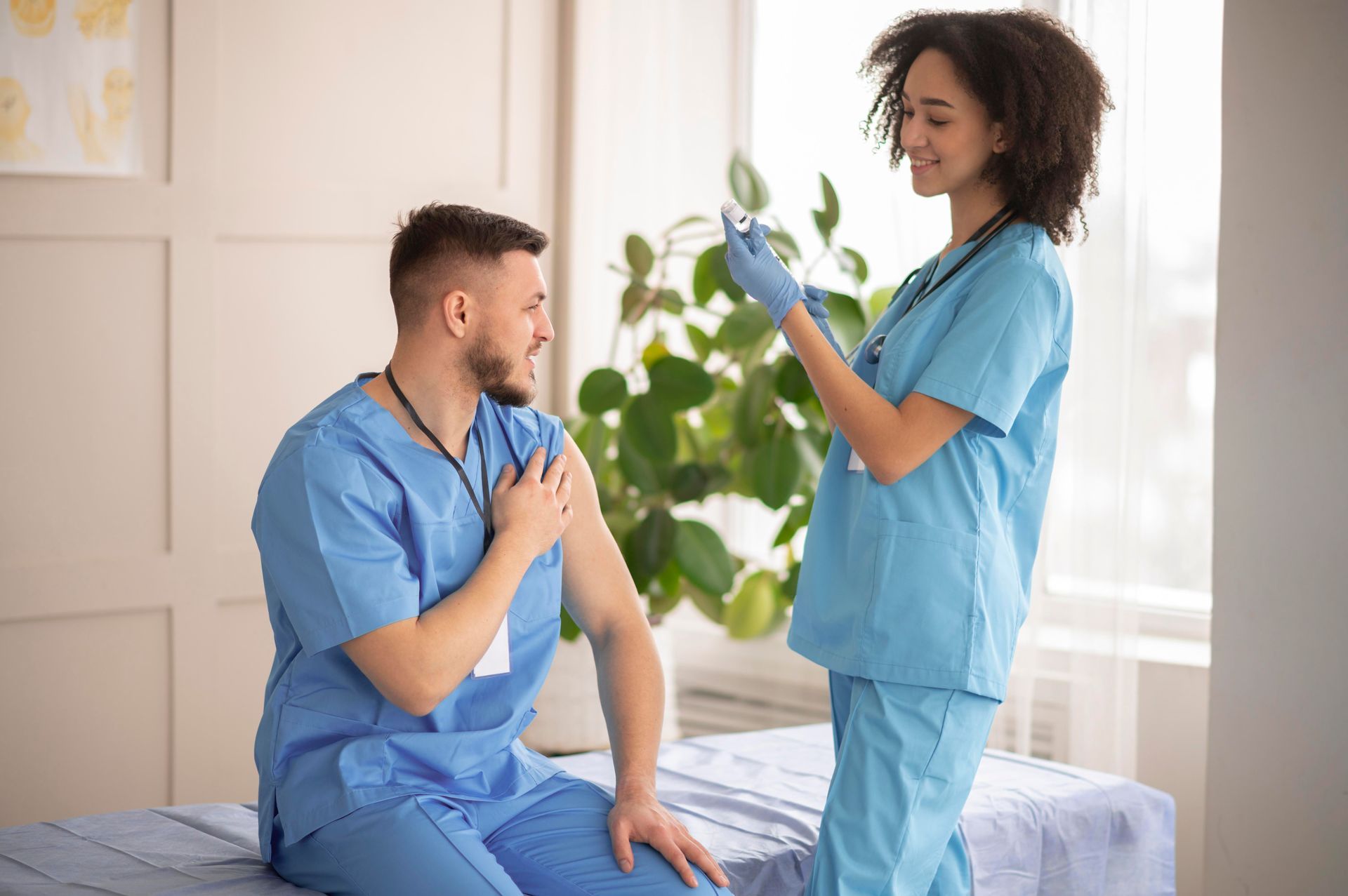 Elderly person sitting, writing on a notepad. Caregiver in background folds clothes in a bright room.