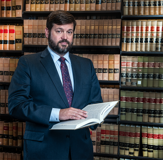Man in suit, holding open book, in front of a bookcase filled with books.