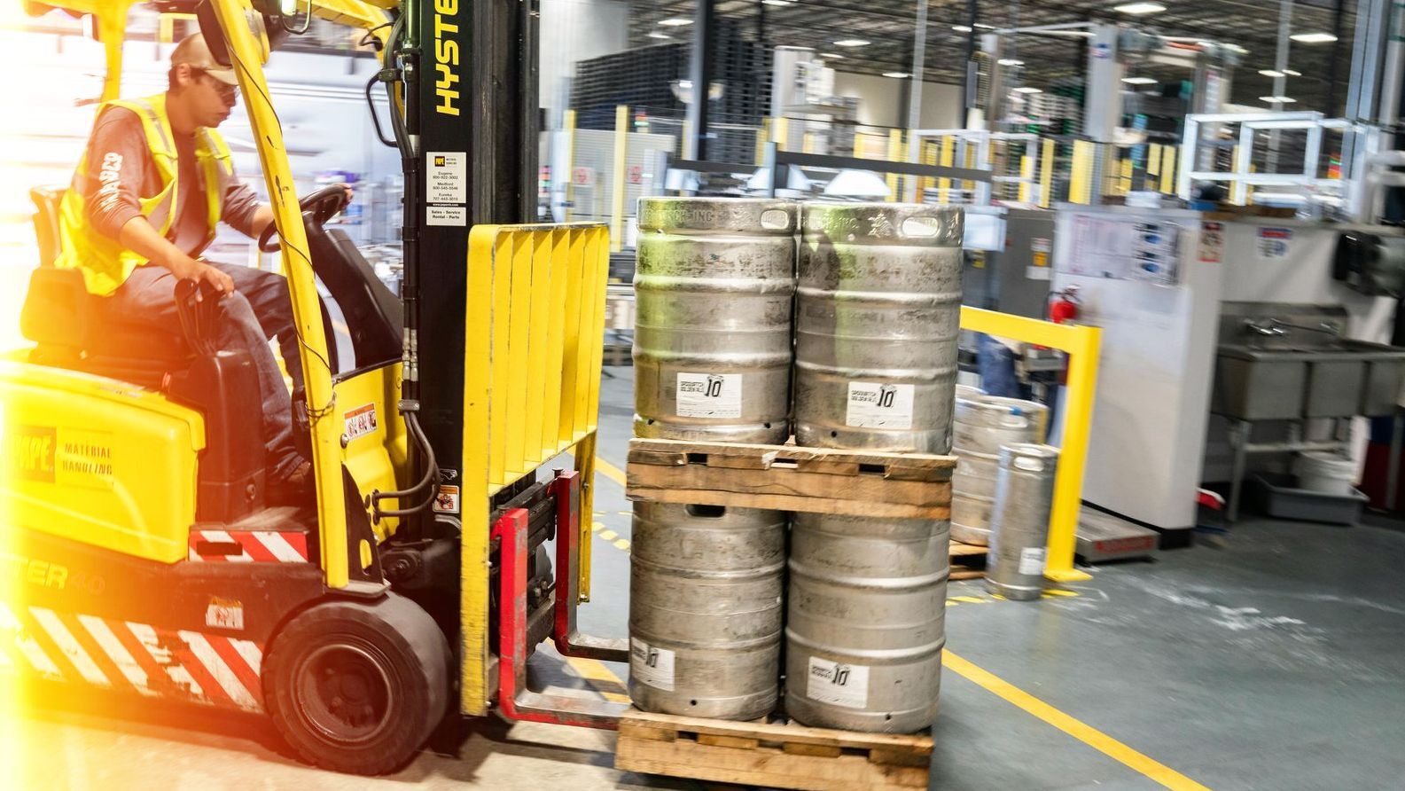 A man is driving a forklift in a factory carrying a pallet of kegs.