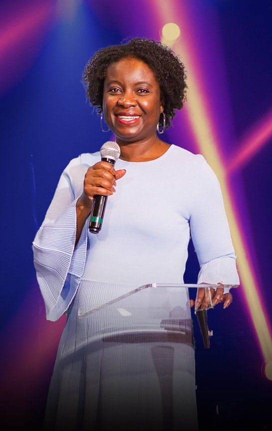 Pastor Grace is standing at a podium holding a microphone and smiling.