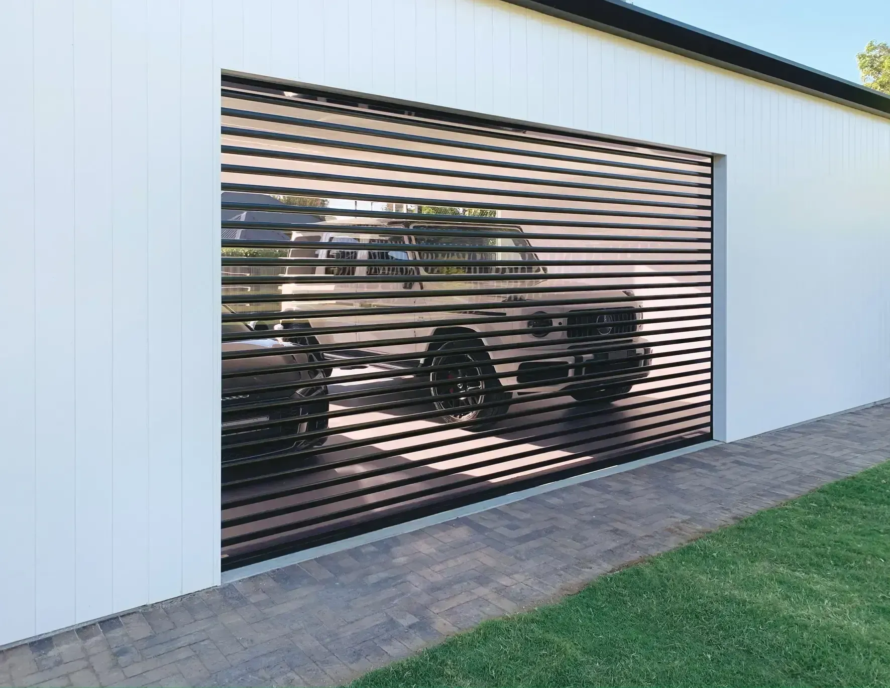 Garage with a car inside, visible through a horizontal slat-style door. White building with brick walkway and green grass.