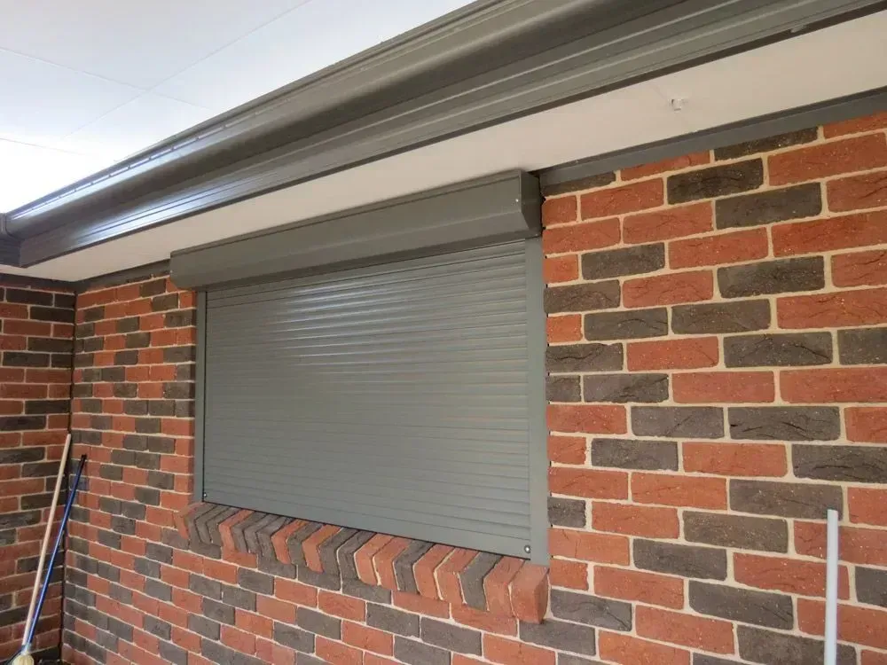 Gray rolling shutter over a window on a brick building, under a gray roof.