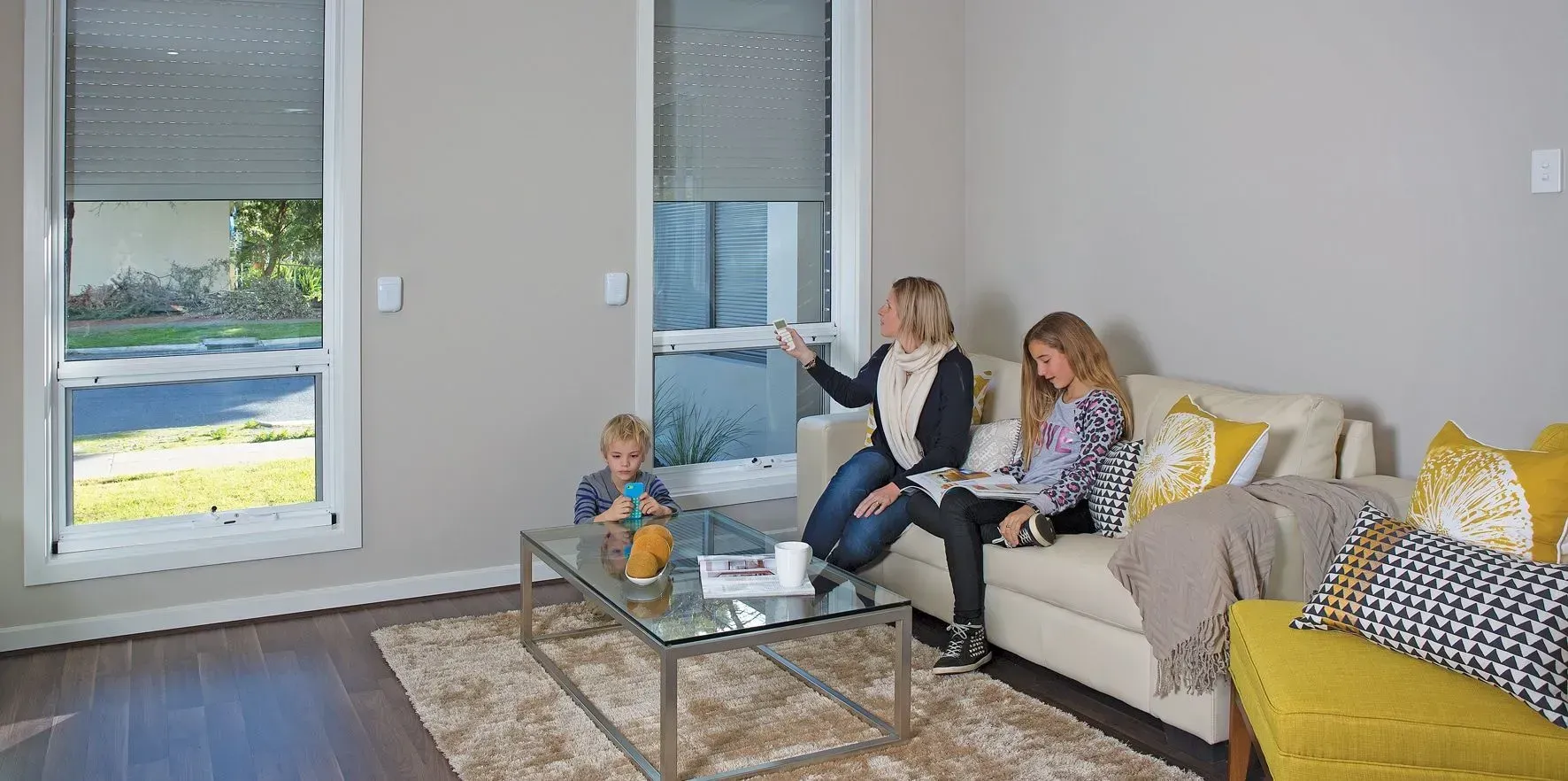 A family sitting in a living room with a couch, coffee table, and windows.