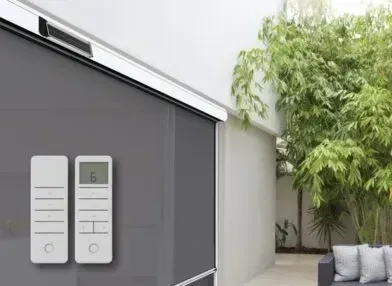 Gray automated outdoor blinds with a remote control. Blinds are partially lowered, with a bamboo garden in the background.