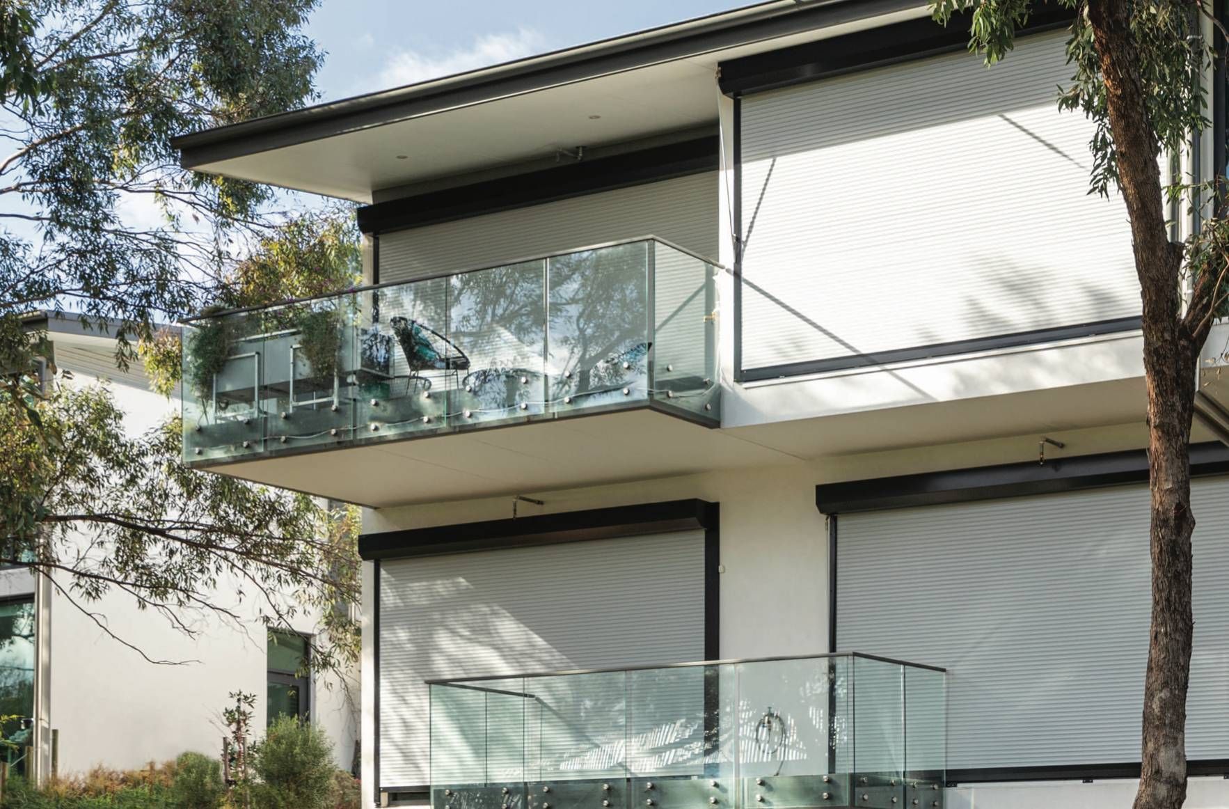 Modern house with glass balconies, gray sunshades, and surrounding trees.