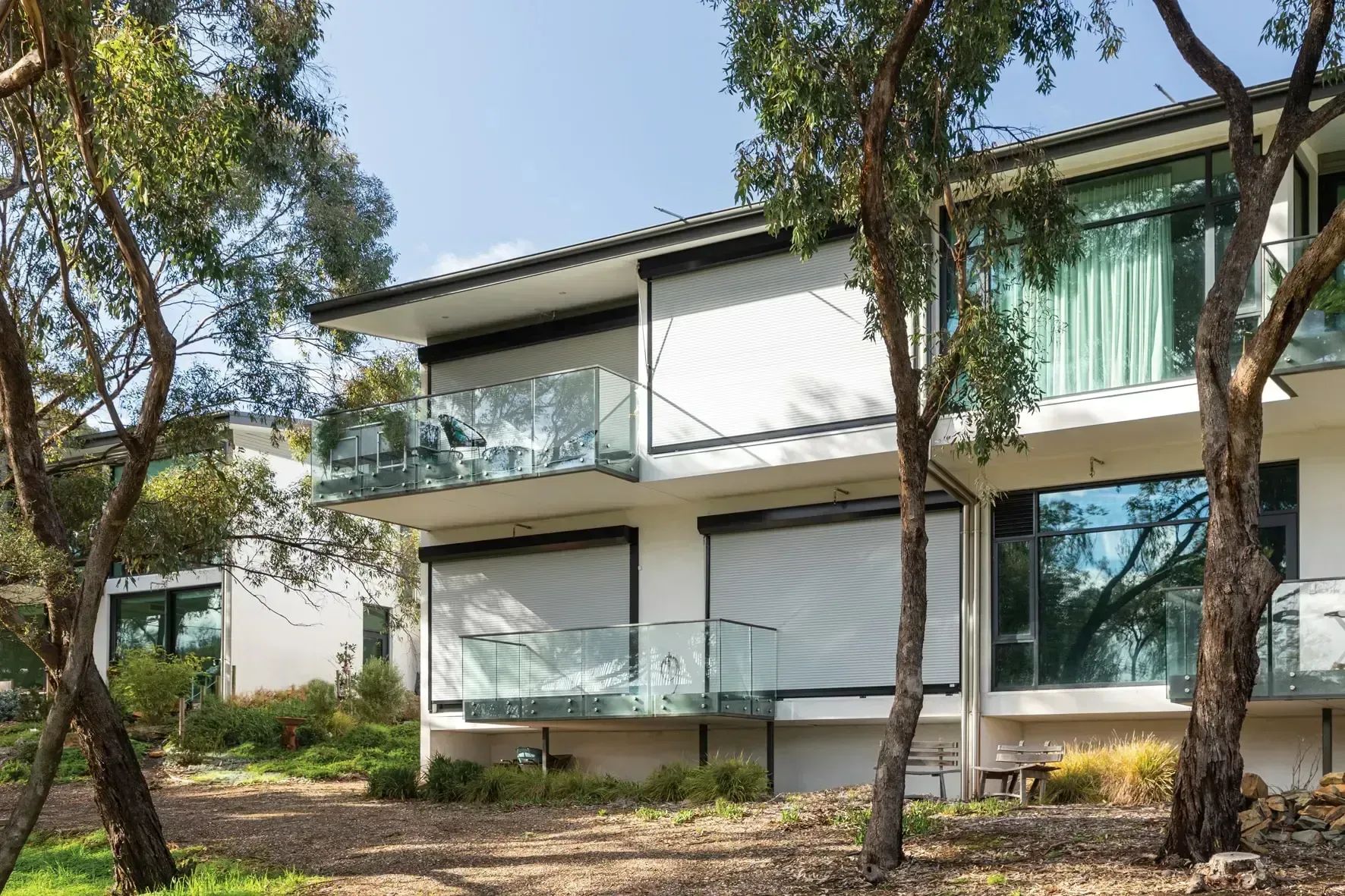 White two-story building with balconies, glass railings, and large windows, shaded by trees.