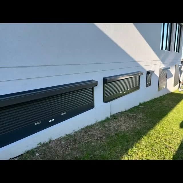 Exterior of a building with black roller shutters over windows, on a gray wall, with green grass.