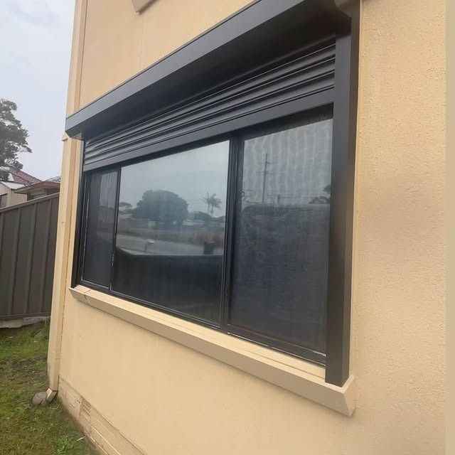 Black roller shutter over a window on a beige house exterior.