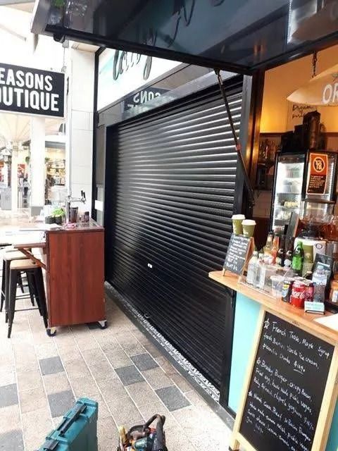 A closed black security shutter on a cafe entrance, with tables and a menu board in view.