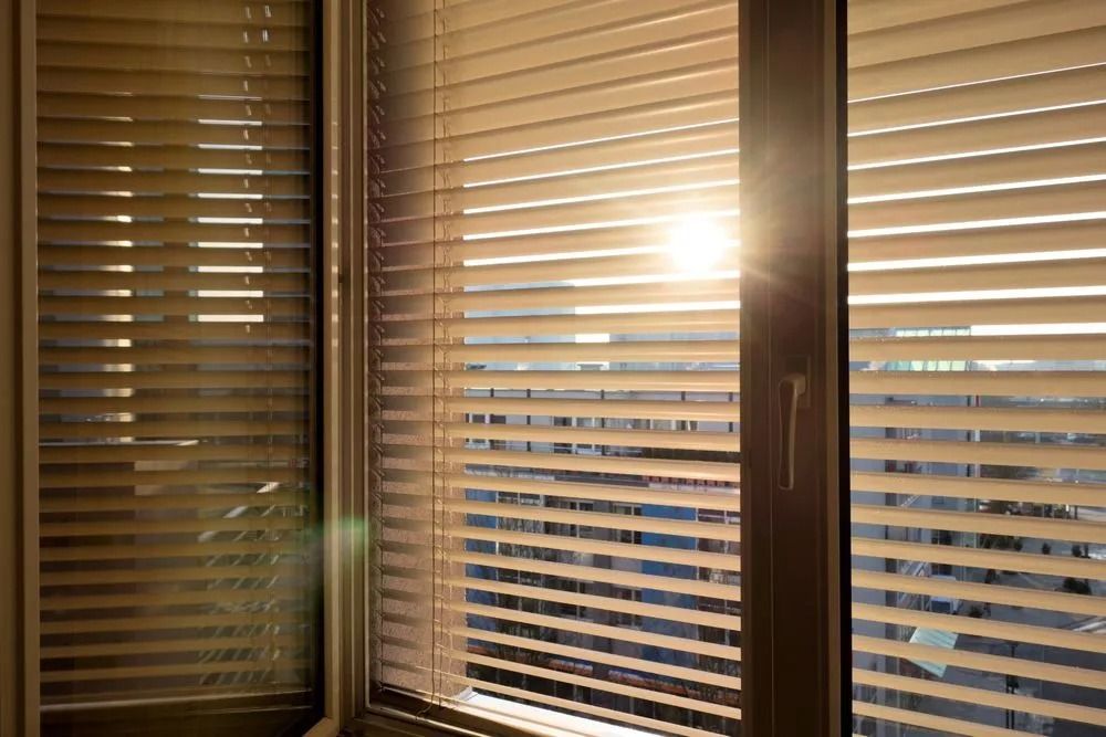 Sunlight streams through beige venetian blinds on a window, casting shadows.
